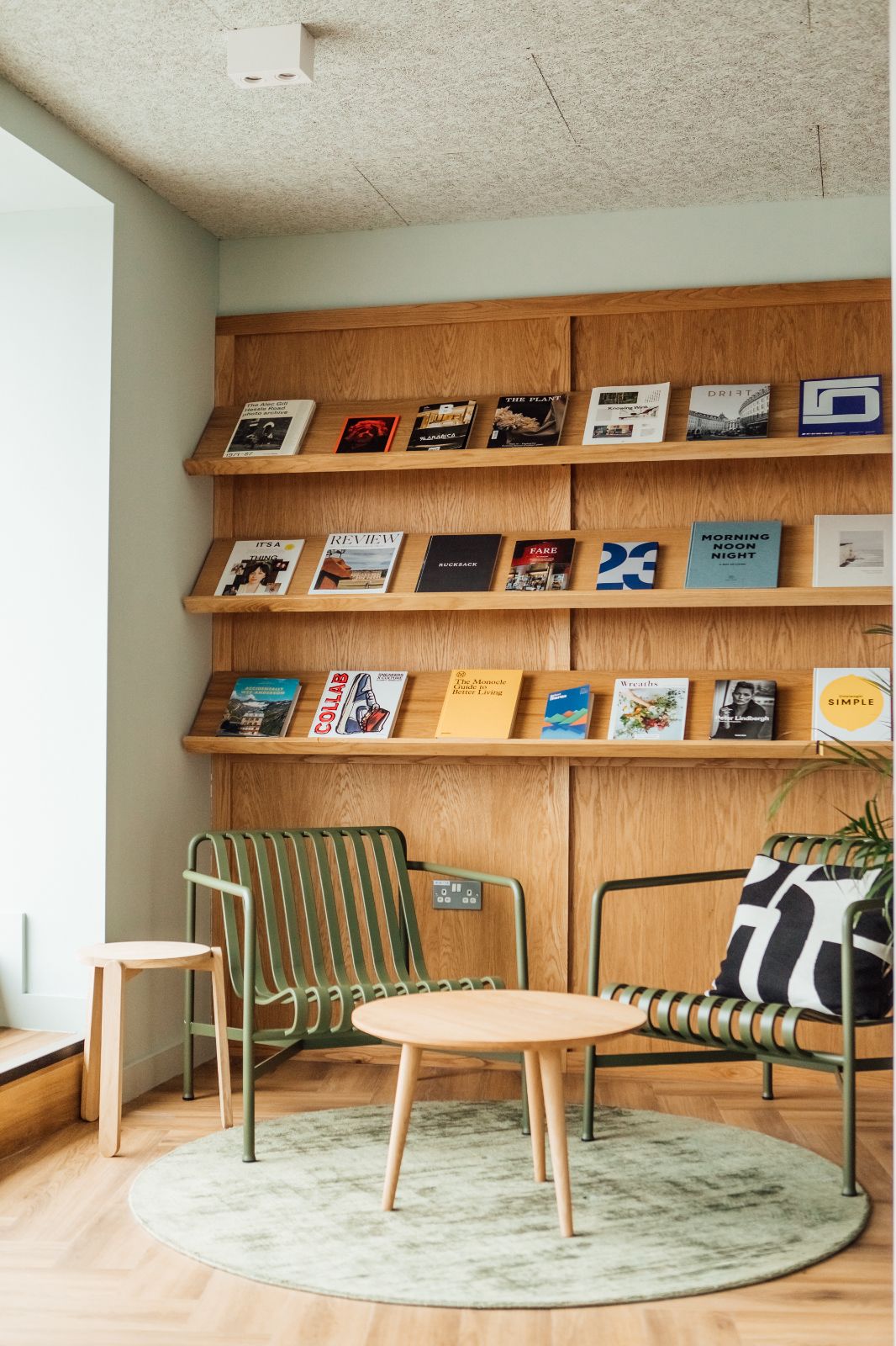 Interior design by 93. Green chairs, sage green circular rug, wooden cladding on the walls with thin bookshelves.